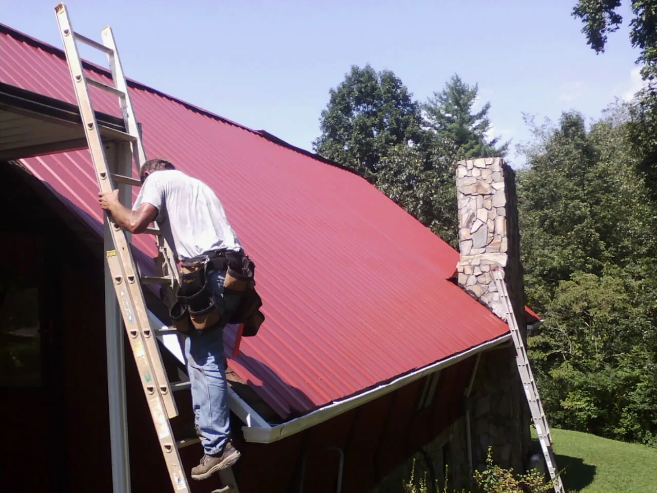 Roofer on a ladder beside a red metal roof and stone chimney — Soffit & Fascia Repair by Roofing Brothers in Cortland